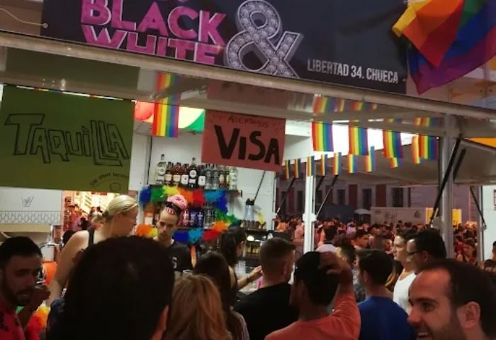 Madrid's Black & White gay club, Chueca, during Pride. Rainbow flags adorn a bar accepting Visa. Crowded street scene, gay travel.