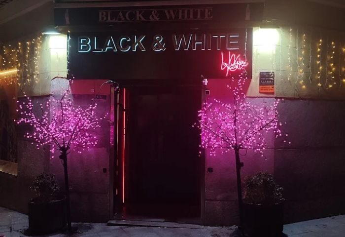 Entrance to Black & White gay club in Madrid. Neon sign, pink trees, and festive lights. Gay travel advice, nightlife hotspot.
