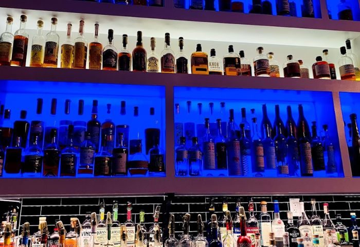 Blackbook drink selection. A bar with many bottles of alcohol on shelves, lit with blue lights, offering a variety of drinks.