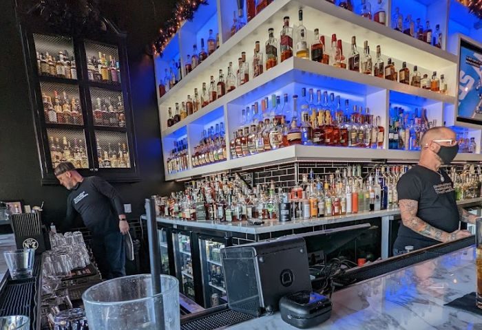 Blackbook drinks: Two bartenders work behind a well-stocked bar with illuminated shelves. A gay bar scene with a wide selection of alcohol.