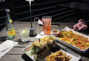 Blackbook Food: Delicious food at a gay bar. Sandwich, fries, nachos, and drinks on a wooden table. Great spot for gay men's travel.