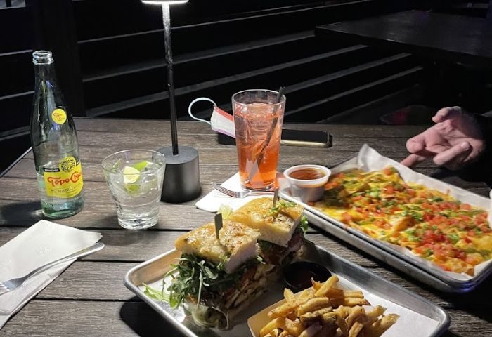 Blackbook Food: Delicious food at a gay bar. Sandwich, fries, nachos, and drinks on a wooden table. Great spot for gay men's travel.