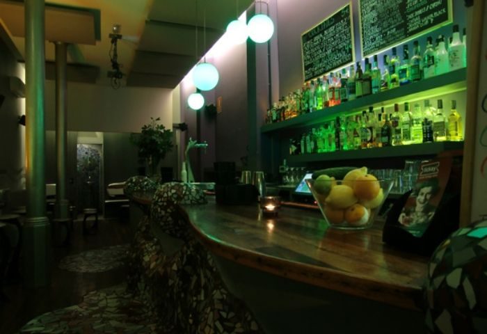 Interior of Blunit Gay Bar in Barcelona. Bottles line shelves under soft green lighting. A wooden bar is in the foreground.