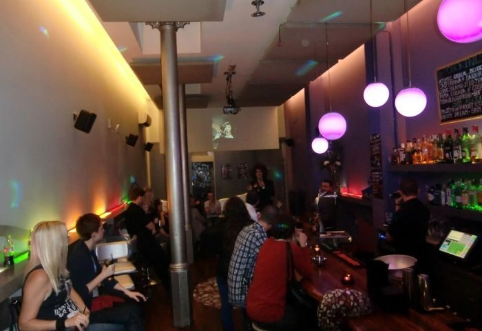 Blunit Gay Bar in Barcelona: Interior view of the bar with patrons, bar counter, lighting, and a performance area. Gay travel.