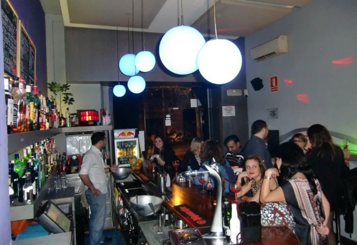 Blunit Gay Bar in Barcelona: Interior view of the bar with patrons, bartenders, and illuminated globe lights. Gay travel advice.