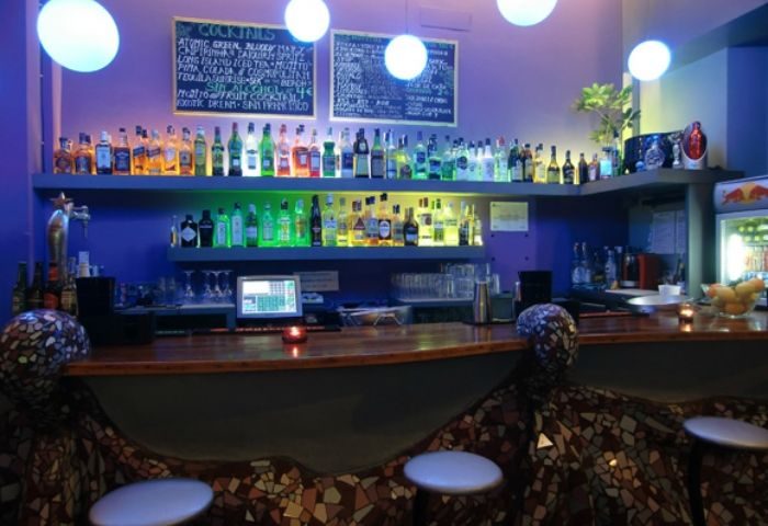 Blunit Gay Bar in Barcelona. Bar counter with mosaic design, shelves of liquor bottles, cocktail menu, and round lights. Gay travel advice.