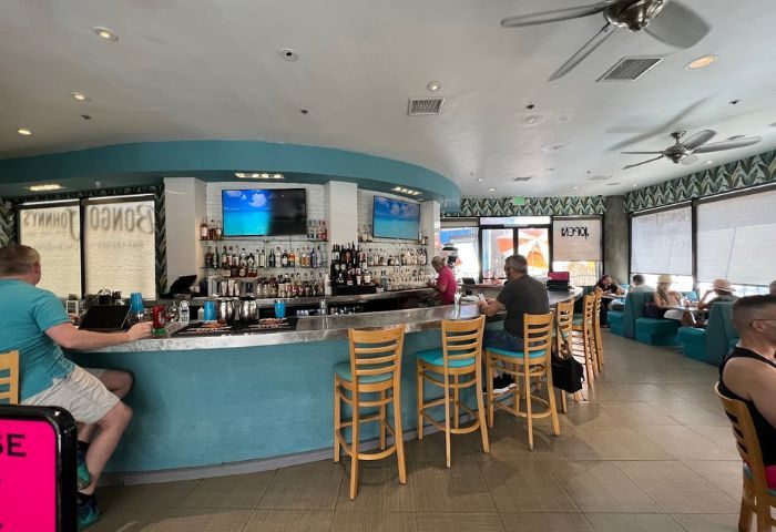 Bongo Johnny's Patio Bar & Grill interior. Bar with stools, liquor bottles, TVs, and patrons. Gay travel destination for drinks and relaxation.