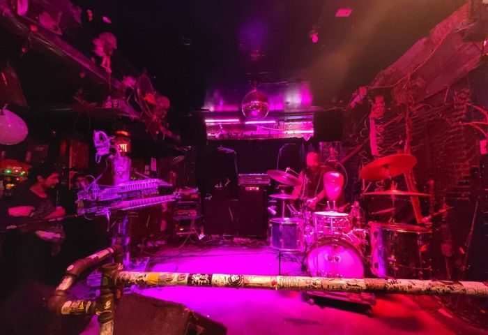 Live music at Bovine Sex Club, Toronto. Band on stage with drums, keyboard, and guitar under pink lighting. Gay travel nightlife.
