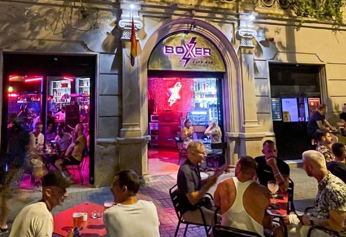 Boxer Cafe Bar, a gay bar in Barcelona. Men sit at tables outside the bar. Rainbow flag hangs near the entrance. Nightlife scene.
