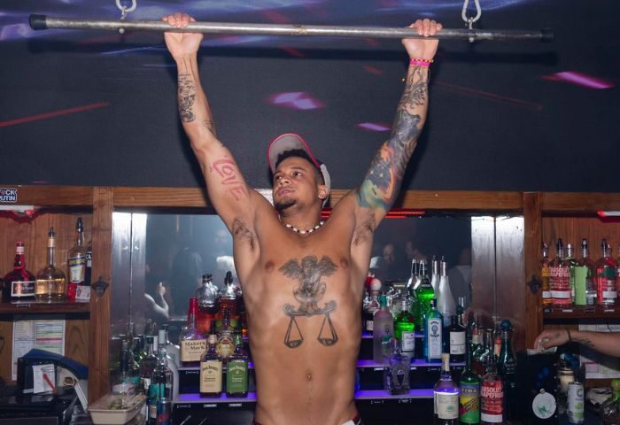Boxers NYC gay sports bar: Shirtless bartender with tattoos hanging from bar in New York. Gay nightlife and travel.