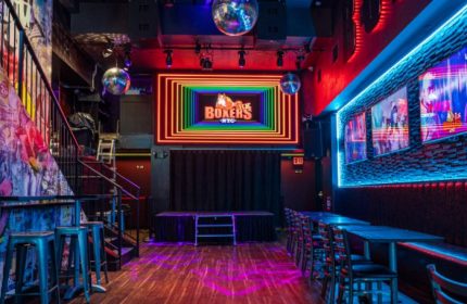 Boxers NYC gay bar interior. Stage, tables, and rainbow neon sign. Gay travel destination in New York City. Nightlife spot for gay men.