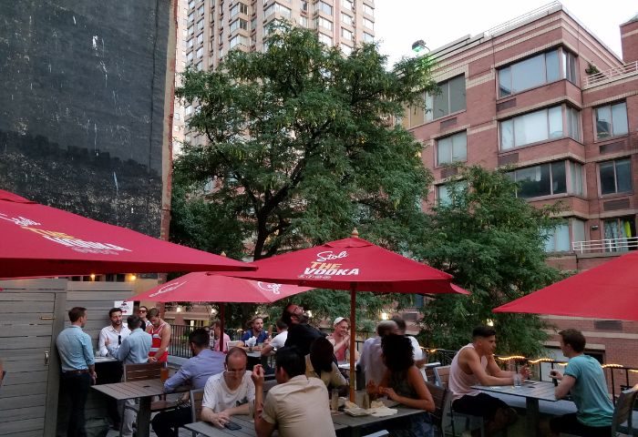 Boxers NYC HK in New York: Outdoor patio scene with gay men at tables under red umbrellas. Urban setting, travel advice.