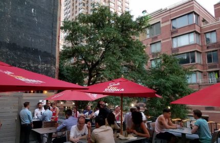 Boxers NYC HK in New York: Outdoor patio scene with gay men at tables under red umbrellas. Urban setting, travel advice.