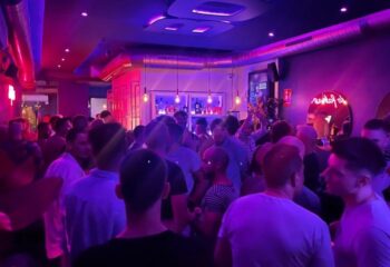 Crowded Boys Bar BCN, a gay bar in Barcelona, features a lively atmosphere with men socializing under vibrant purple and pink lighting.