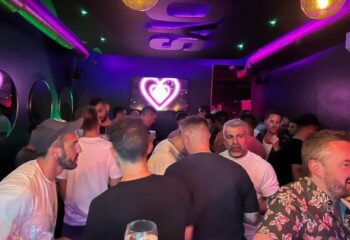 Crowded Boys Bar BCN, a gay bar in Barcelona. Purple lighting, heart-shaped neon sign, and diverse group of men socializing. Gay travel.