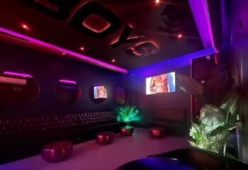 Interior of Boys Bar BCN, a gay bar in Barcelona. Features include purple lighting, leather seating, and screens showing male torsos.