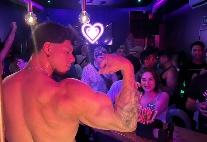 Shirtless man flexing at Boys Bar, a gay bar in Barcelona, Spain. Heart-shaped neon sign in background. Gay nightlife travel.