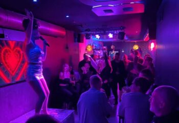 Gay bar in Sitges: Drag queen performs on stage at a boys' bar. Crowd enjoys drinks and entertainment in a vibrant, purple-lit atmosphere.