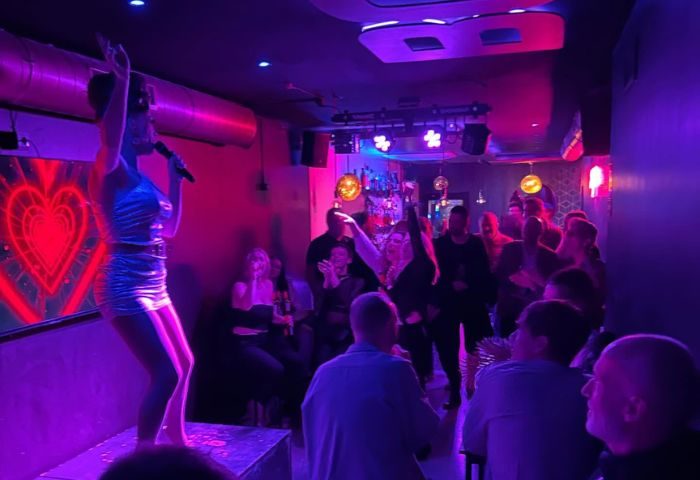 Gay bar in Sitges: Drag queen performs on stage at a boys' bar. Crowd enjoys drinks and entertainment in a vibrant, purple-lit atmosphere.