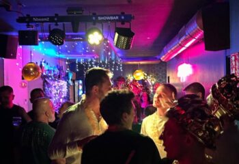 Gay bar scene in Sitges. Men enjoy drinks in a crowded bar with colorful lights and festive decorations. Gay travel nightlife.