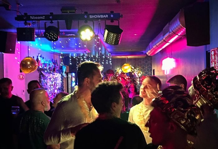 Gay bar scene in Sitges. Men enjoy drinks in a crowded bar with colorful lights and festive decorations. Gay travel nightlife.