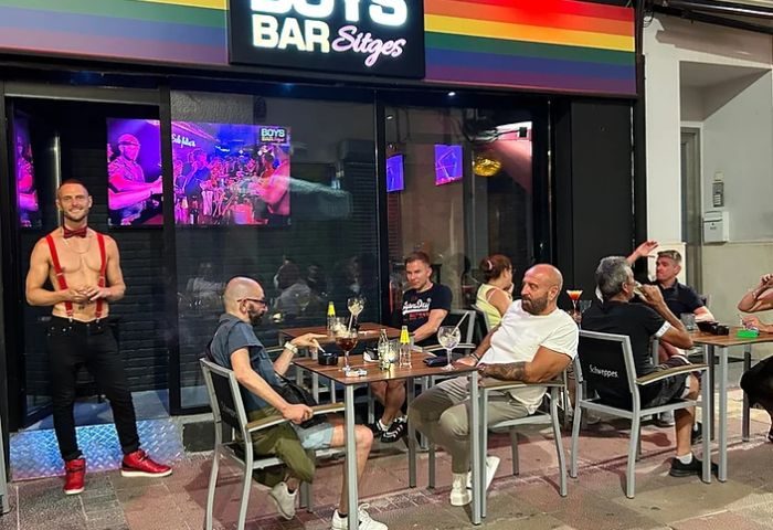 Boys Bar Sitges, a gay bar, features men enjoying drinks outside. Rainbow flag above entrance. A shirtless man in suspenders stands nearby.