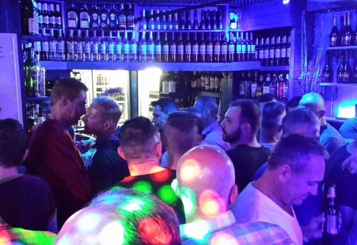 Busy gay bar in Benidorm. Men gather under blue lights, drinks line shelves. Nightlife scene, Brief Encounter gay venue. Travel advice.