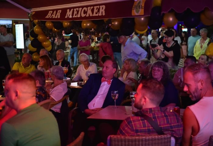 Patrons at Buddies Bar, a gay bar in Gran Canaria, enjoy drinks. The bar's awning reads Bar Meicker. Balloons decorate the scene.