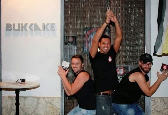 Three men pose at Bukkake Cruise Club, a gay cruise club in Sitges. They're smiling and holding cards. Gay travel advice.