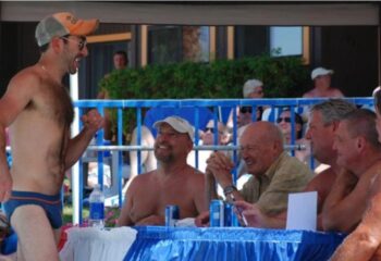 CCBC Resort Hotel Gay Cruise Palm Springs event: A man in swimwear celebrates, with judges at a table and onlookers behind.