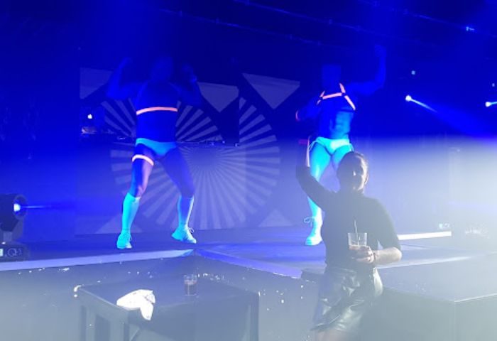 CENTURYON Gay Club in Torremolinos: Two dancers in UV gear perform on stage. A person with a drink watches. Nightlife, gay travel.