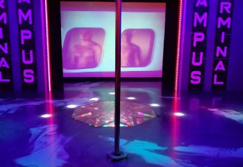 Interior of Campus, a gay strip bar in Montreal. A chrome pole stands center stage, with screens showing shirtless men. Neon lights add to the ambiance.