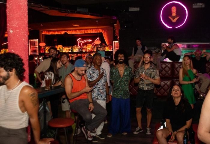Candy Darling gay bar in Barcelona. A group of gay men & women socialize in a dark bar with neon lights and a well-stocked bar.