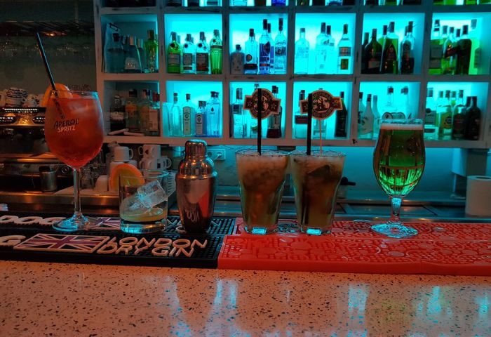 Central Bar, a gay bar in Sitges. Cocktails on the bar with a shelf of bottles behind, illuminated by blue lights. Gay travel.