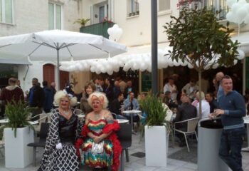 Central Bar Books, a gay bar in Sitges. Drag queens pose at an outdoor seating area. People gather under white balloons.