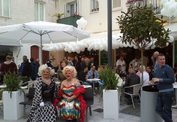 Central Bar Books, a gay bar in Sitges. Drag queens pose at an outdoor seating area. People gather under white balloons.