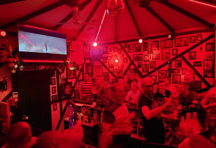 Centre Stage Bar, a gay bar in Gran Canaria. Red lighting, crowded with men. Photos on the walls, disco ball, and TV screen.