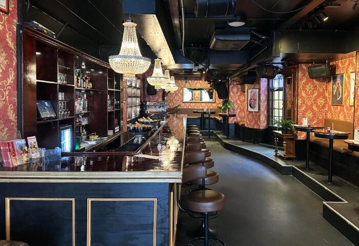 Centre Stage CPH gay bar interior. Chandeliers hang over a long bar with stools. Red patterned walls, tables, and booths line the raised back.