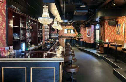 Centre Stage CPH gay bar interior. Chandeliers hang over a long bar with stools. Red patterned walls, tables, and booths line the raised back.