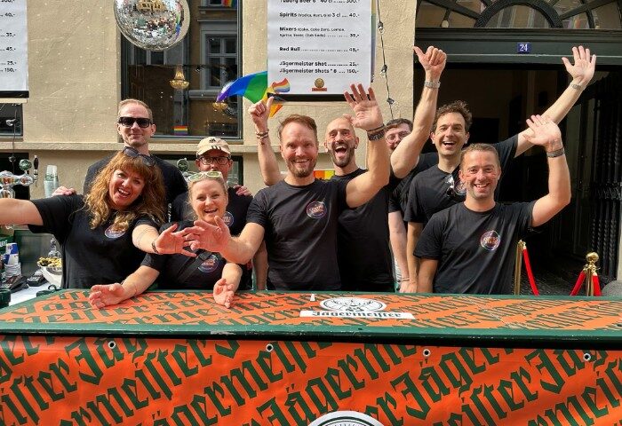 Centre Stage CPH: Gay bar staff in Copenhagen waving. Disco ball, rainbow flag, and Jägermeister bar. Gay travel advice, nightlife.