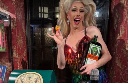 Drag queen at Centre Stage CPH, a gay bar in Copenhagen. She's holding a Jägermeister bottle and a numbered ball, smiling.