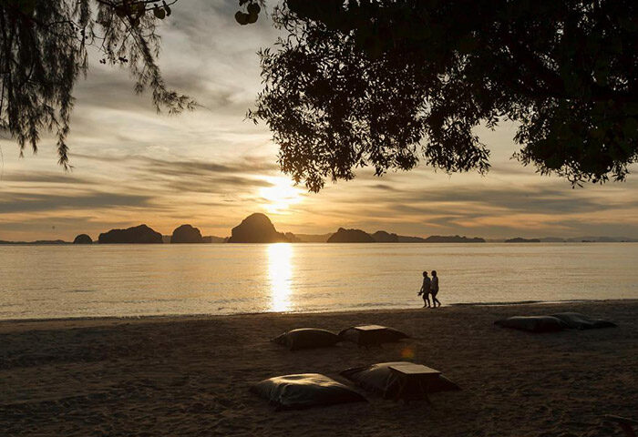 Sunset at The Tubkaak Krabi Boutique Resort, a beachfront luxury hotel. Two people walk along the beach, with islands in the distance. Krabi, Thailand.