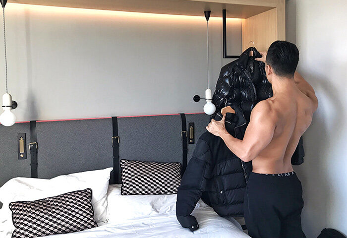 Gay-friendly Assembly Hotel, London. Shirtless man with black pants hanging up a jacket. Modern, cheap hotel room with unique headboard design.