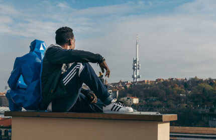 Man at meetme23, a gay hostel in Prague near Vinohrady, sits with a blue statue overlooking the city and Žižkov Television Tower. Gay nightlife.