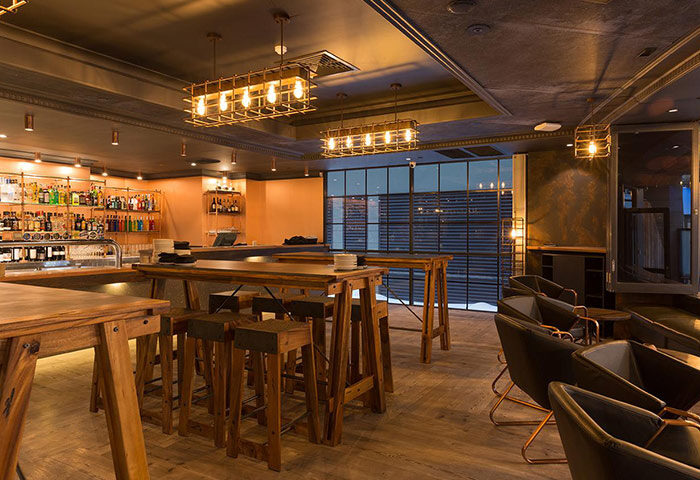 Rydges World Square hotel bar in Sydney's Darlinghurst gayborhood. Rustic wood tables, stools, and caged light fixtures create a warm, inviting space.