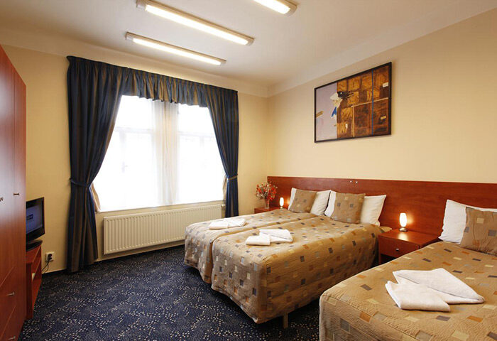 Cheap hotels for three gay men in Prague's Vinohrady gayborhood. Room with twin beds, blue curtains, and patterned carpet. Comfortable stay.