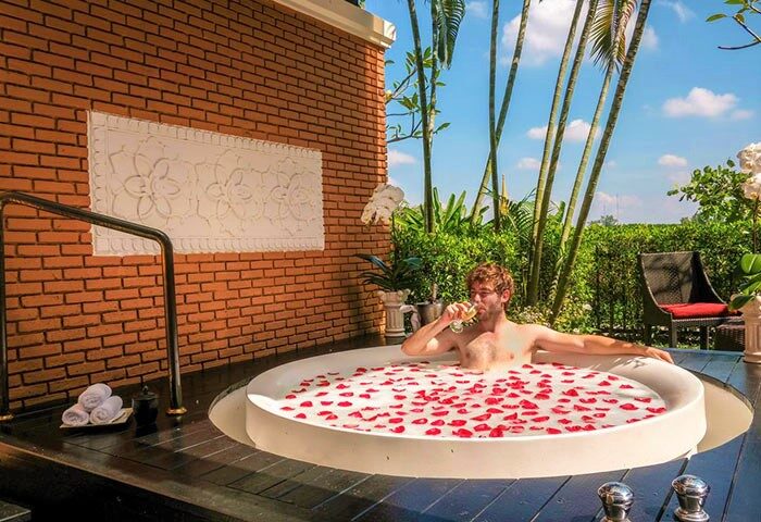Man in luxury Shangri-La Hotel Chiang Mai hot tub with rose petals, drinking wine. Gay travel goals in Thailand. Cheap LGBT vacation.