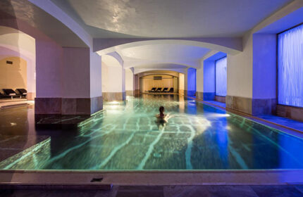 Indoor pool at NH Collection Carlo IV, a luxury gay hotel in Prague's Old Town. Person swimming, columns, arches, and lounge chairs visible.