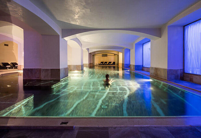 Indoor pool at NH Collection Carlo IV, a luxury gay hotel in Prague's Old Town. Person swimming, columns, arches, and lounge chairs visible.