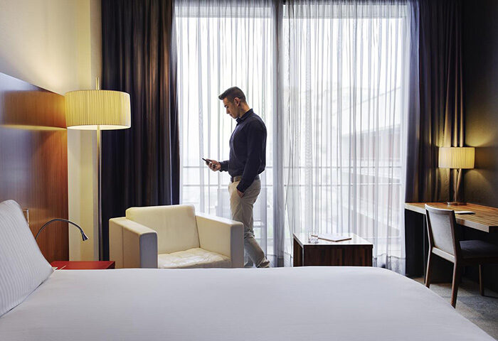 Man in Sofitel Barcelona Skipper hotel room. Gay-friendly hotel near Mar Bella gay beach. Modern interior, desk, chair, and sheer curtains.